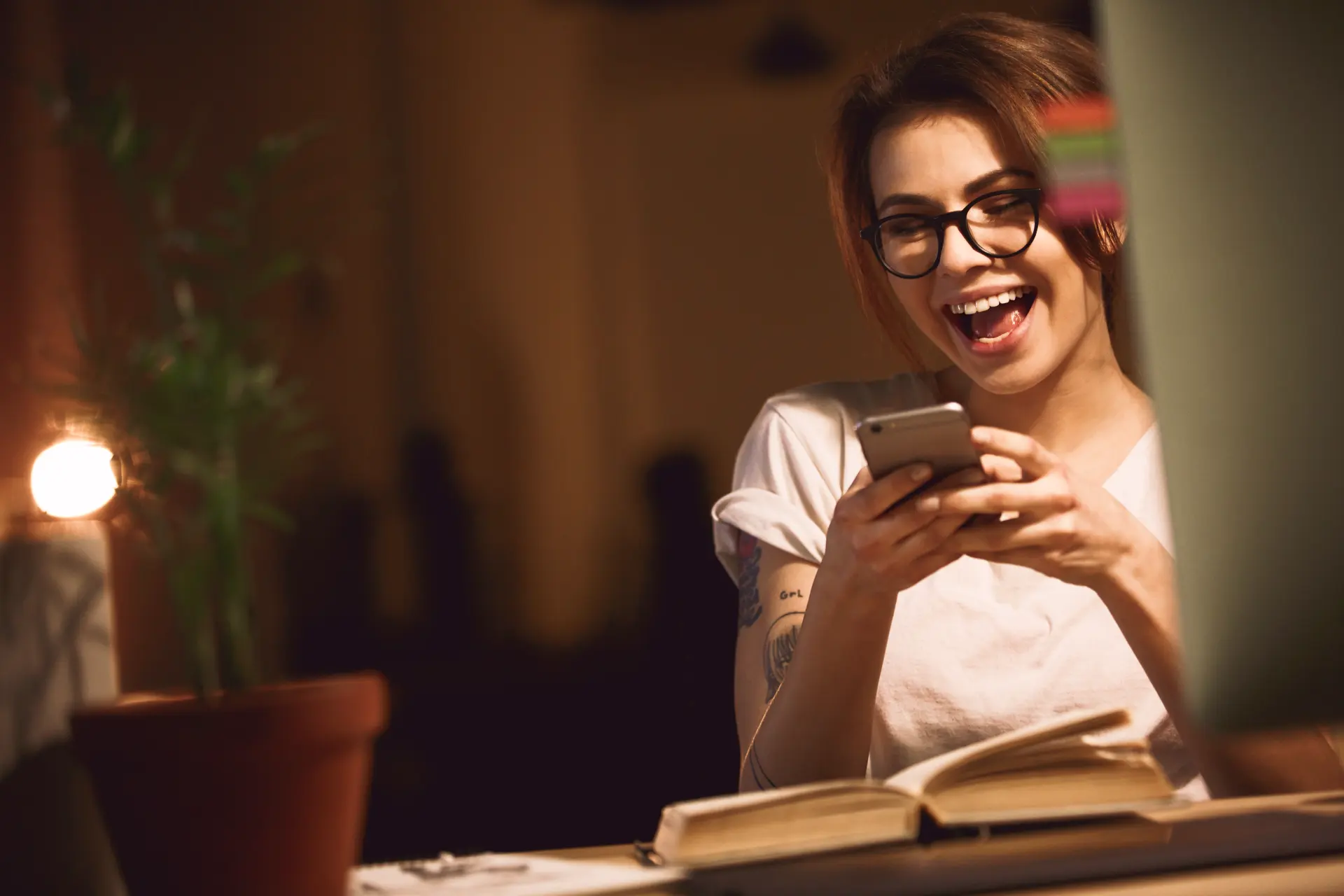 Mulher sorrindo enquanto lê uma conversa no celular, à noite, numa mesa com planta e luminária