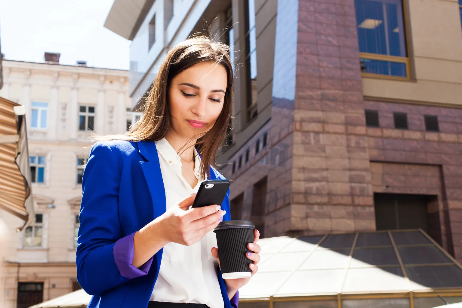 Mulher lendo uma mensagem no celular na rua, com um copo de café na mão